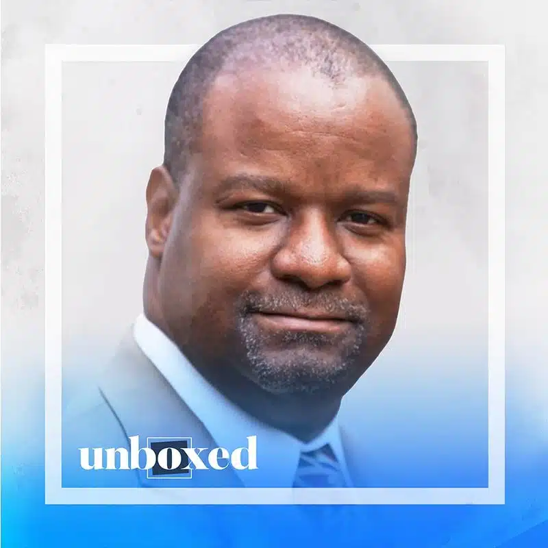 A person wearing a suit and tie is smiling at the camera, exuding a sense of nurture. The image has a gradient blue overlay at the bottom with the word unboxed in white text. The background is plain and light-colored, reflecting calm amidst challenging times.