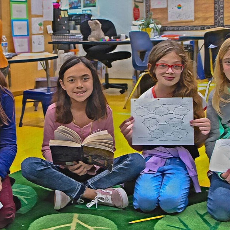 Four children are sitting on a green carpet in a classroom, their voices animated as they discuss the open books and paper with diagrams. The background shows chairs and colorful educational materials, creating an inviting exhibition of curiosity. They appear engaged and happy.