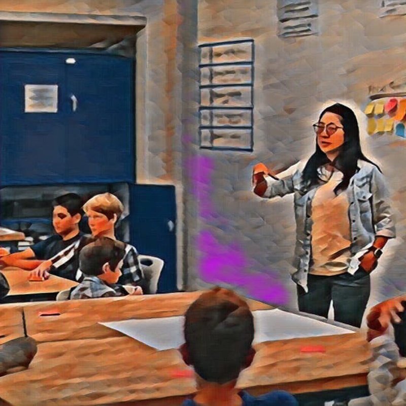 A teacher stands at the front of a classroom, engaging students seated at tables. The students are listening attentively. The image has a painted effect with prominent purple and blue hues.