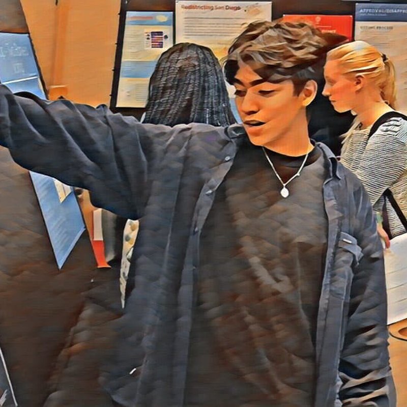 A person with dark hair and a black shirt gestures while standing in front of display boards, likely explaining a project. Another person with long hair focuses on the boards, and a blonde-haired individual stands nearby. The setting appears to be an exhibition or presentation space.