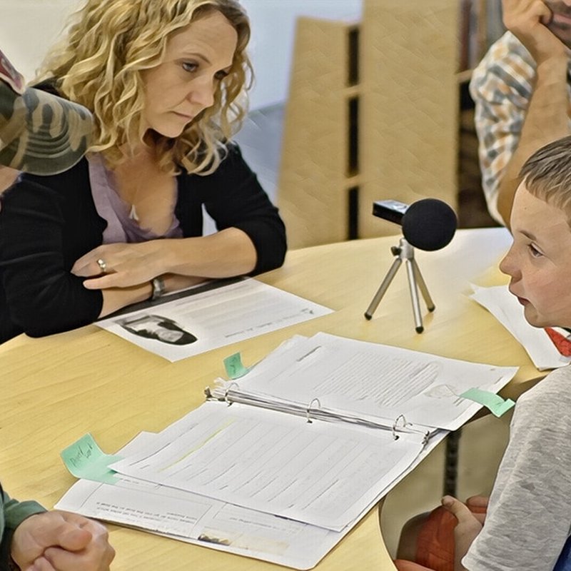 A woman attentively listens to a boy speaking at a table, which has papers, a binder, and a microphone. The setting suggests student-led conferences or a discussion. Two other people are partially visible in the background, emphasizing the collaborative atmosphere.