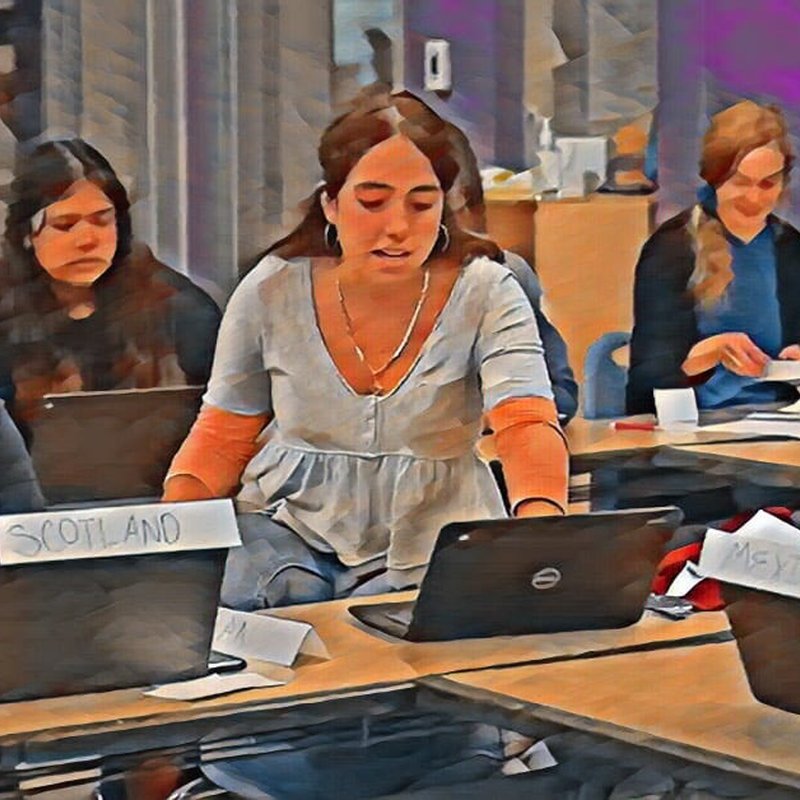 A group of people sits at a table with laptops, engaged in discussion. It resembles a PBL setting or classroom. Nameplates, including one labeled Scotland, are visible on the table. The image has an artistic, painted effect, embodying the creative spirit of a Design Kit approach.