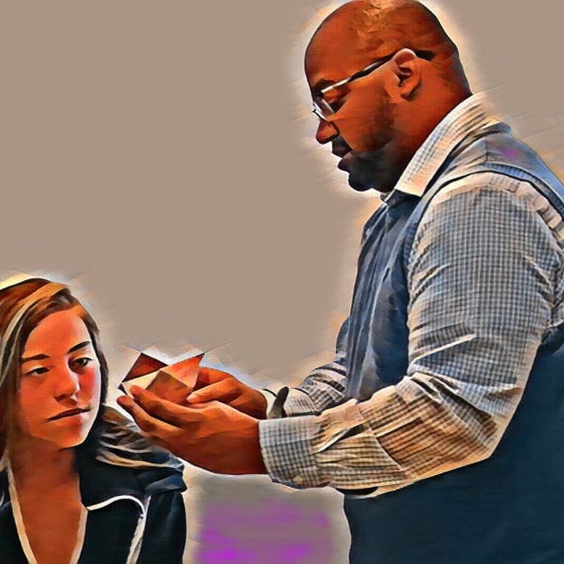 A man in glasses and a collared shirt holds a red and white geometric object from a Design Kit, explaining its cycles of reflection to a woman with long hair, who is attentively listening. The background is neutral, and the image has an artistic filter effect.