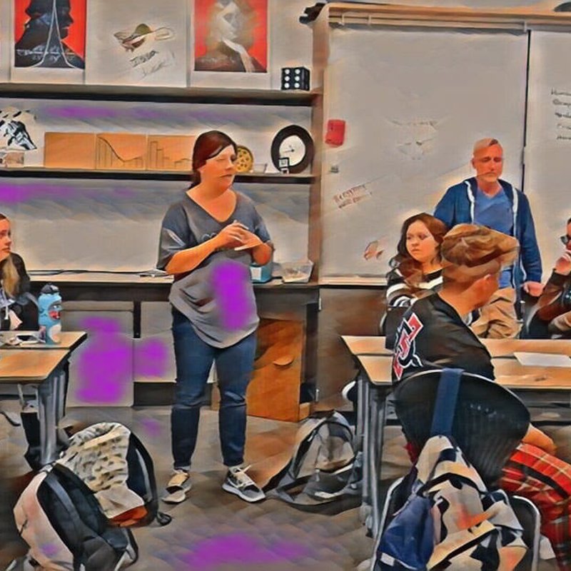 A teacher stands in front of a classroom, fostering educational excellence as students listen attentively at their desks. Books and posters adorn the walls, while backpacks rest on the floor, creating an environment geared toward great academics in this high school setting.