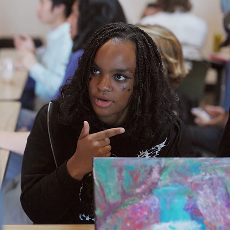 A person with braided hair and face markings gestures while sitting at a table, engaged in a lively conversation about student-centered assessment. Surrounded by peers, they delve into the colorful painting before them as part of their education evaluation project.