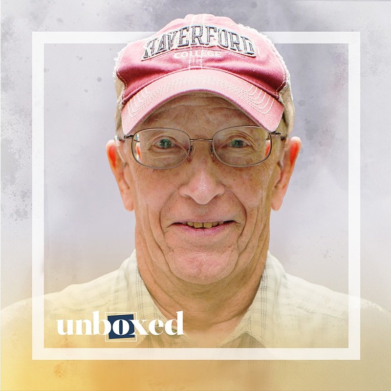 A smiling older adult, exuding wisdom, wears glasses and a red cap that reads Haverford College. They are framed by a white border with unboxed in blue and yellow at the bottom. The light, slightly textured background adds to the serene aura of this captivating scene.