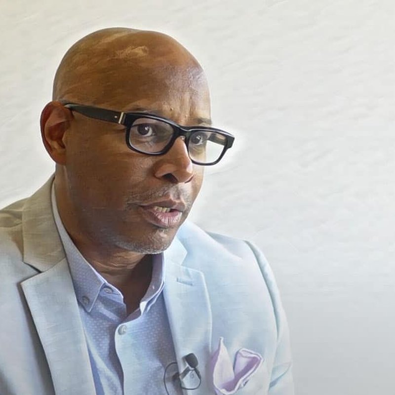 A person in a light gray suit and glasses is speaking, honoring the dedication of students. The background is plain and light-colored, reflecting an atmosphere focused on education.
