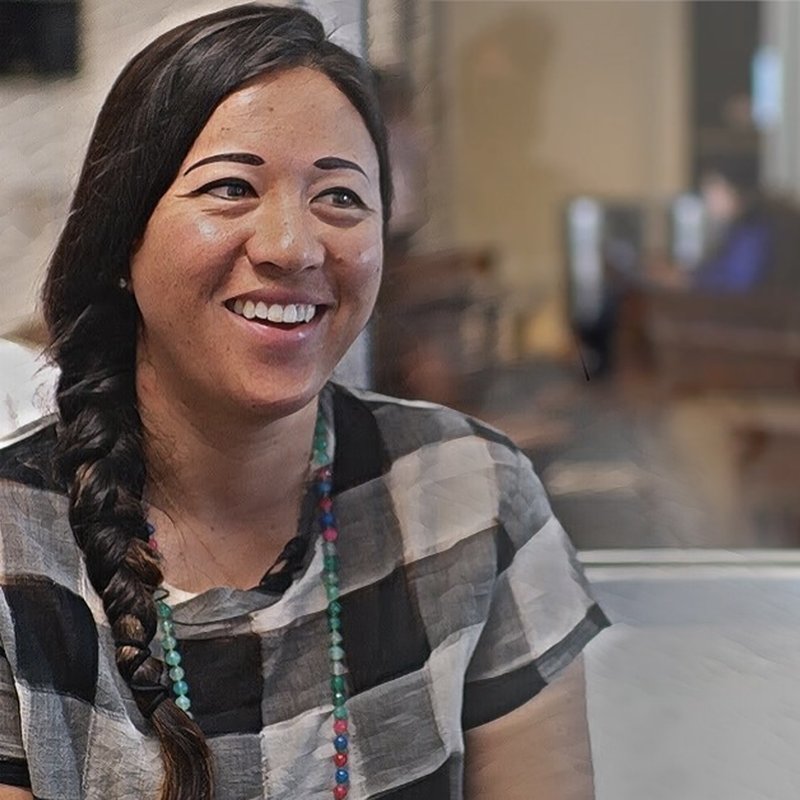 A person with long, dark hair in a braid is smiling. They are wearing a black and white checkered shirt and a colorful beaded necklace, embodying the spirit of authentic projects. The background is softly blurred, showing an indoor setting with people and furniture.
