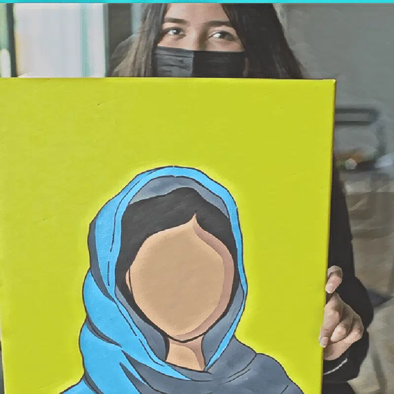 A person at an exhibition holds a painting of a faceless figure in a blue hijab. Theyre partially visible, wearing a black mask and sporting dark hair, embodying the Power of the Influencer through art.