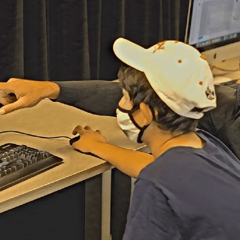 A young person in a cap and mask sits at a desk, focused on a computer screen. An adults hand points at the monitor, guiding them with a fun project. The scene is set against curtains and computer equipment.