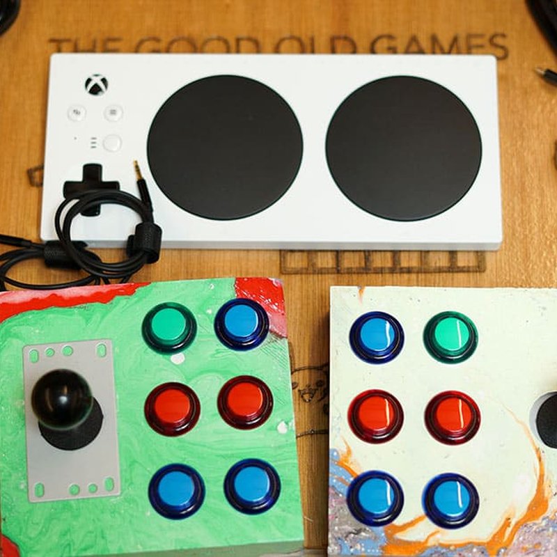 AbleGamerz adaptive gaming controllers are displayed on a wooden surface in this project overview. The top device features a white base with two large black circular buttons, while below sit two colorful controllers embellished with blue, red, and green buttons.