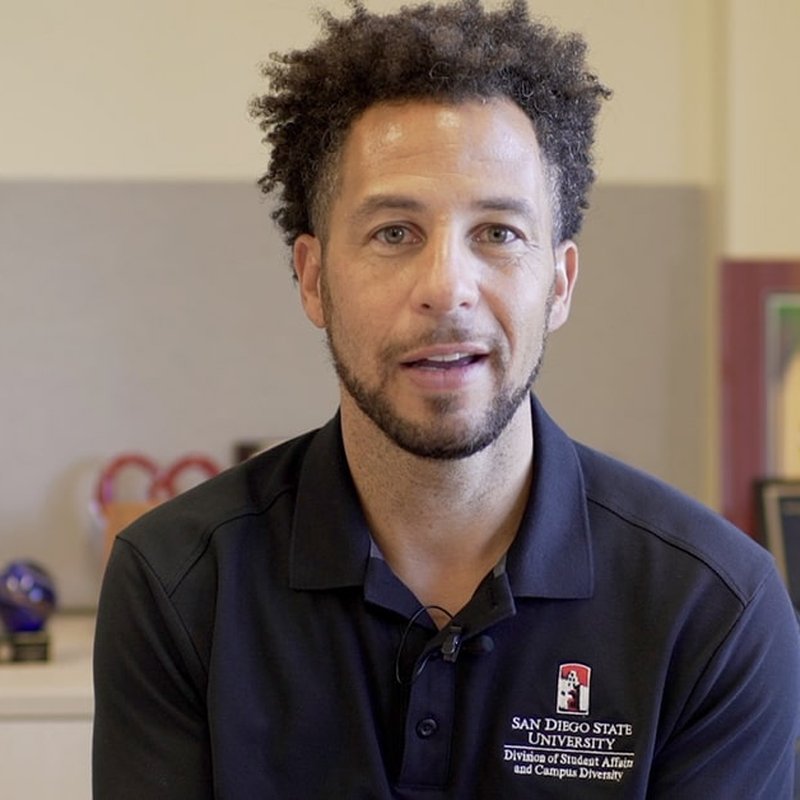 A person with short curly hair, wearing a navy blue polo shirt with the San Diego State University logo, sits indoors. The background is softly blurred, showcasing office decor and awards, a testament to achievements not just by them but also by others.