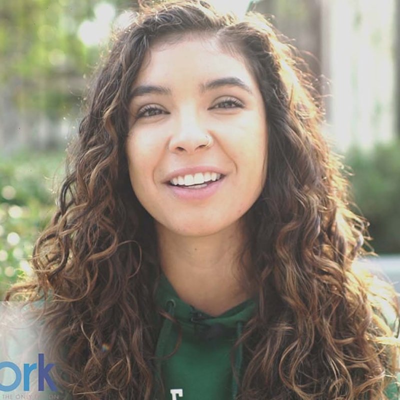 A person with long curly hair and a nose ring smiles at the camera, wearing a green hoodie that aligns with Lemon Grove Academy Middles spirit. The softly blurred greenery in the background complements their connection to the CARE Network’s nurturing environment.