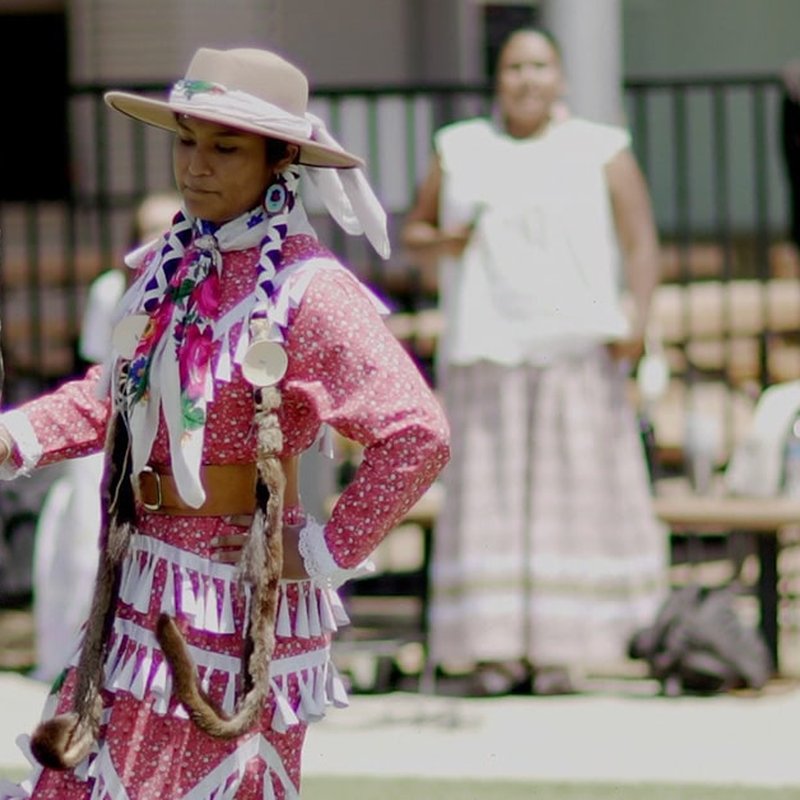 A person dressed in a colorful, traditional outfit with a hat and ribbons performs outdoors like the vibrant Bird Dancers. Another person in the background wears a light-colored dress. They are in a sunlit area with a black fence behind them, reminiscent of Project Deepening’s vivid cultural showcases.