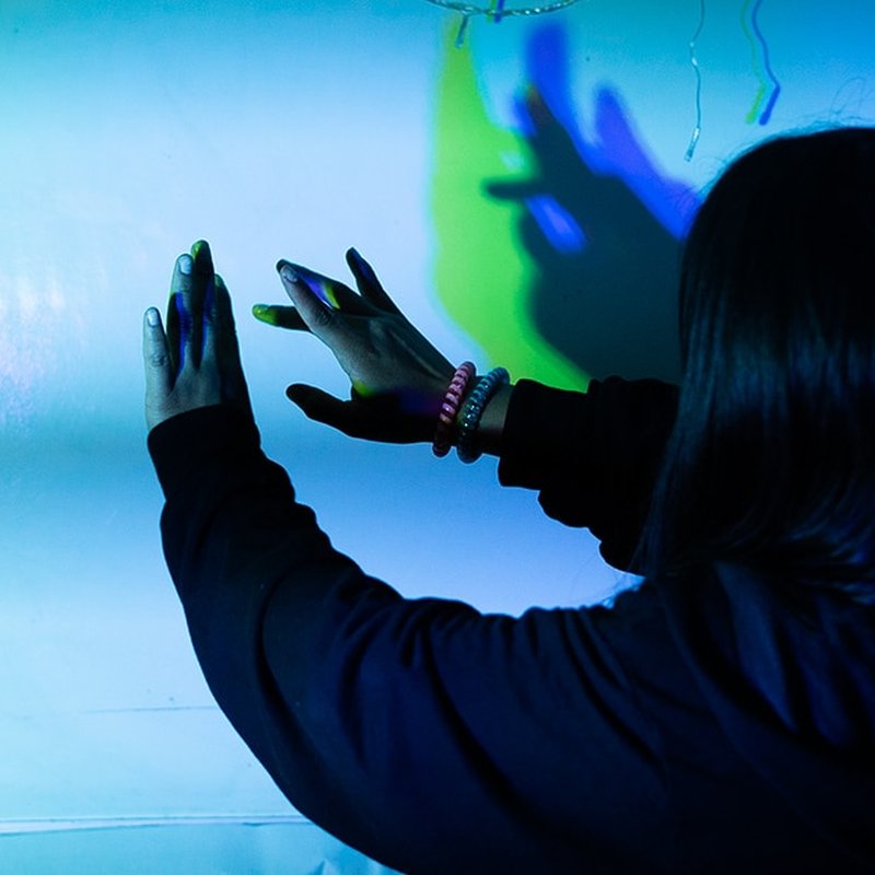 Amidst a mesmerizing play of blue and green light, a person with long hair extends their hands toward a wall, creating colorful shadows. Wearing a dark long-sleeved top and bracelets on one wrist, they embody the impact of an artful exhibition in motion.