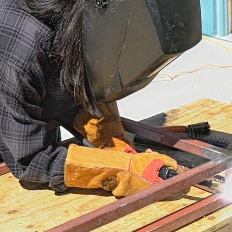 A person is welding metal on a wooden platform, wearing a protective helmet and gloves, seamlessly integrating into the cycles of production. Sunlight is visible in the background, casting a warm glow over this meticulous process.