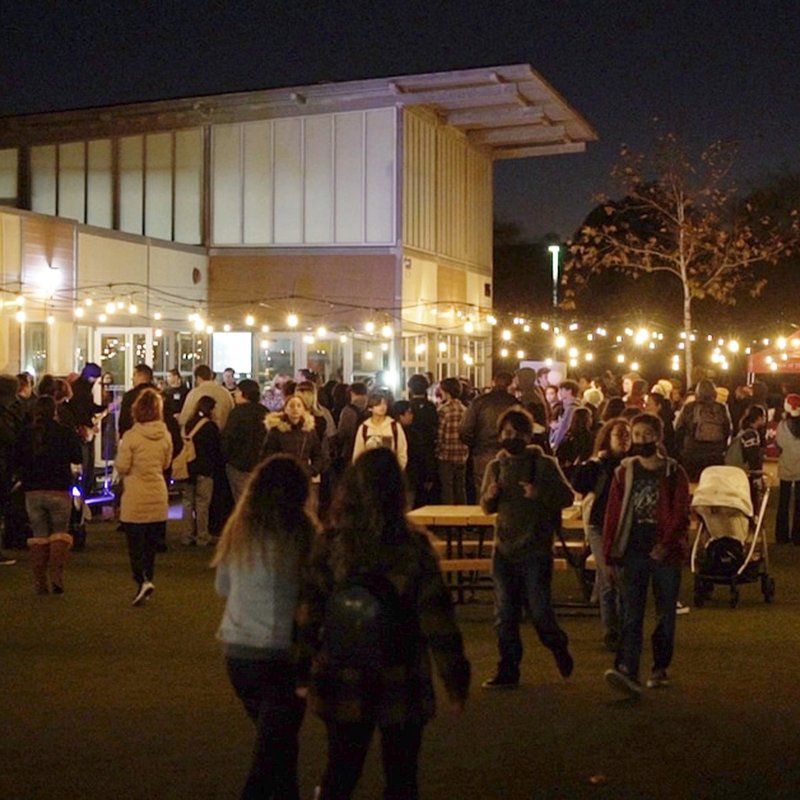 A crowd gathered outdoors at night, with warm string lights illuminating the scene like an open-air exhibition. A modern building is in the background, and people are casually dressed, some with strollers and others engaging in lively conversation or strolling under the cozy illumination.