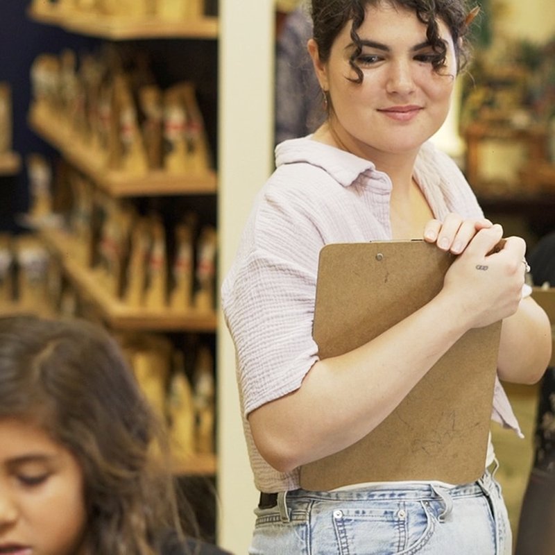 A person stands holding a clipboard, wearing a light-colored shirt and jeans. In the casual, informal setting, shelves brim with items as another person sits at a table nearby. The atmosphere buzzes with collaborative experiences that inspire personalized learning.