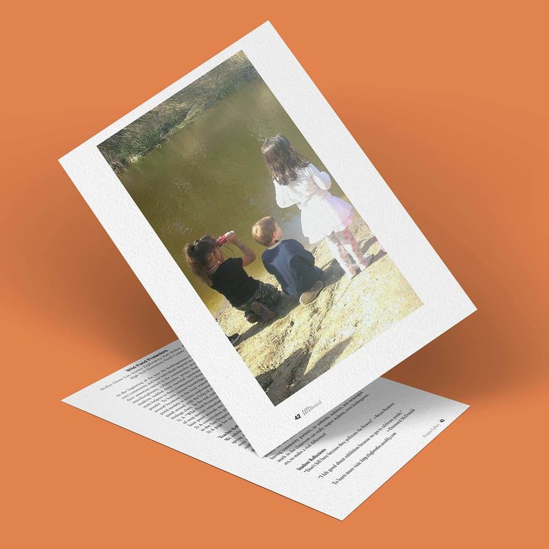 A printed photo captures three children by a wild pond; two are gazing at the water while one sips from a bottle. The image, angled just right, rests on a page of text against an orange background, evoking the quiet vigilance of young protectors in nature.