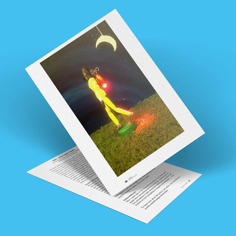 A tilted image on a white card depicts a model figure walking on grass under a crescent moon in a night sky, holding a red lantern. Resembling an ethereal scene from Junk Puppet Theatre, the card rests on a sheet with text against a blue background.