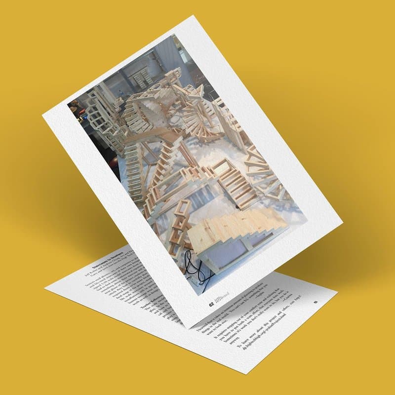 A printed image of an intricate architectural model featuring staircases that seem to lead nowhere, crafted from wood and resting on a page of text. The background is a solid mustard-yellow surface.