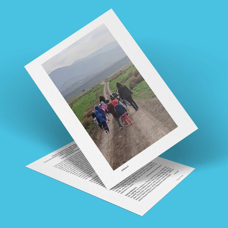 A photo captures a group of people, including children, strolling along a dirt path in a rural area, surrounded by natures treasures. They are dressed in casual clothing. The image leans against another piece of paper on a light blue background.