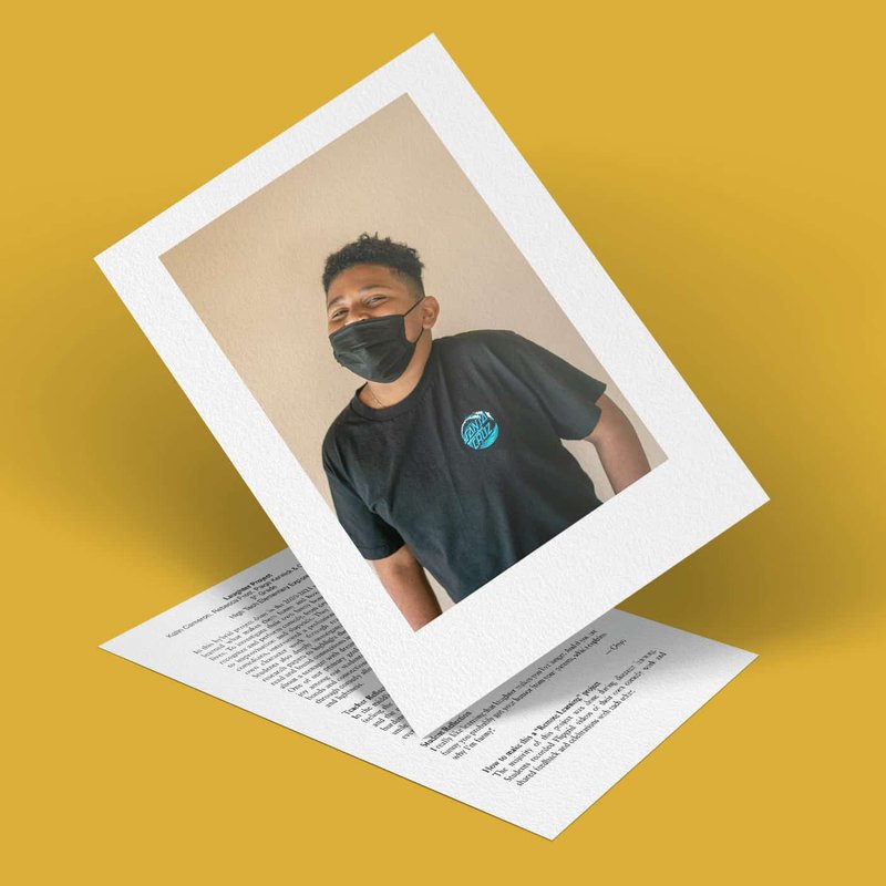 A person wearing a black mask and a black t-shirt with a blue emblem is pictured against a neutral background, exuding an air of mystery. The photo, perhaps part of an intriguing project, rests on a document with printed text, all set on a mustard yellow surface.