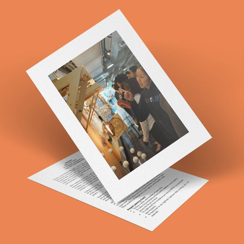 A photo of people observing a display at an event is set on an orange background. The image appears to be printed and is placed atop a piece of paper with visible text, both slightly tilted.