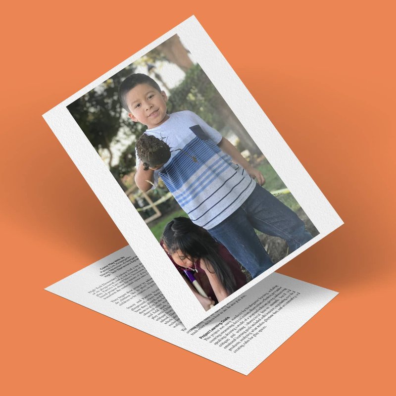 A photo of a smiling child holding a pine cone beckons us to play, propped up on an orange surface. Below the photo is a sheet with printed text. A girl is partially visible, looking downwards. The background shows trees and a fence.