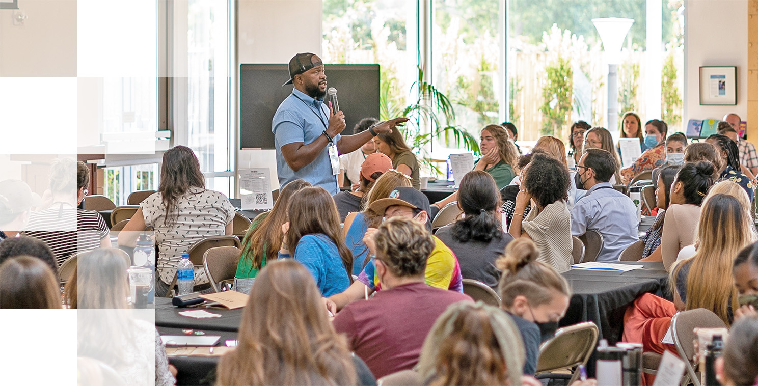 A speaker in a light blue shirt and cap discusses school system leadership with a diverse audience seated at round tables in a bright, modern conference room. Participants are attentively listening and taking notes as large windows illuminate the space.