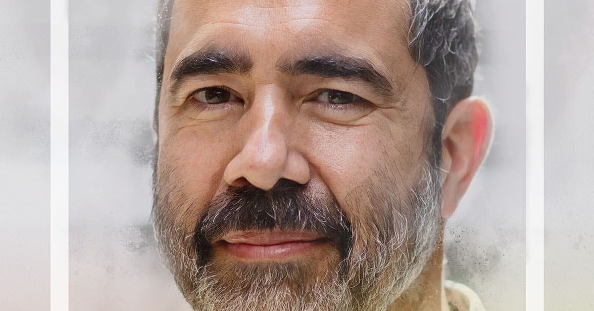 A man with a beard and salt-and-pepper hair, reminiscent of a seasoned educator, is looking at the camera with a neutral expression. The blurred background enhances the focus on his face, as if capturing the wisdom gained through years in education.