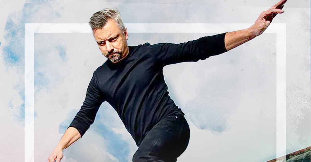 A man in a black long-sleeve shirt and pants appears to be balancing like a skateboarder against a cloudy sky background, his arms extended for stability.