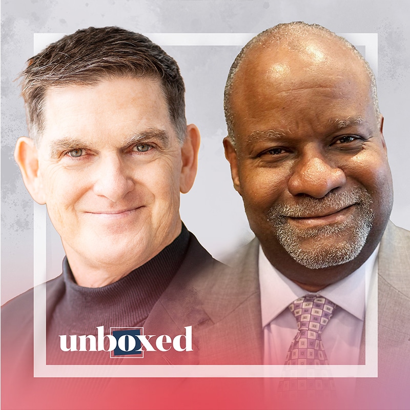 Two smiling men in a professional portrait, one wearing a dark turtleneck and the other in a suit and tie, embody innovation. A pink gradient overlay highlights the word unboxed, capturing Derek Mitchell and Scott Sampsons pioneering spirit in Regenerative Education.