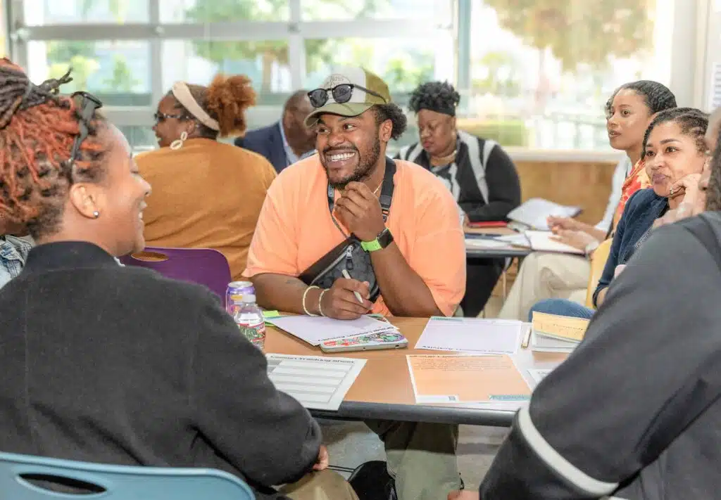 A group of people is sitting around a table, engaged in conversation. At the center, someone is smiling, wearing an orange shirt and cap. Papers and notebooks scatter the table, reflecting a collaborative meeting at the High Tech High Graduate School of Education.