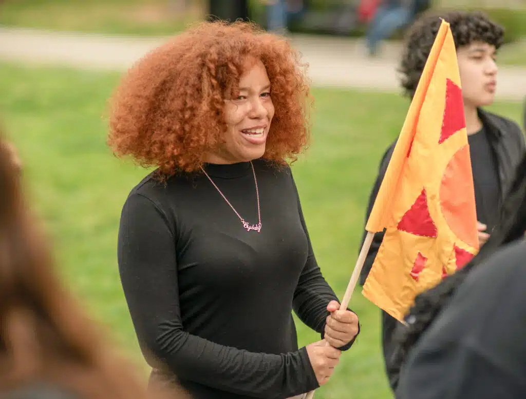 A person with curly red hair, wearing a black outfit, smiles while holding a flag with a yellow and red sun design. They stand outdoors on a grassy area near the Graduate School of Education. Other people are in the blurred background.