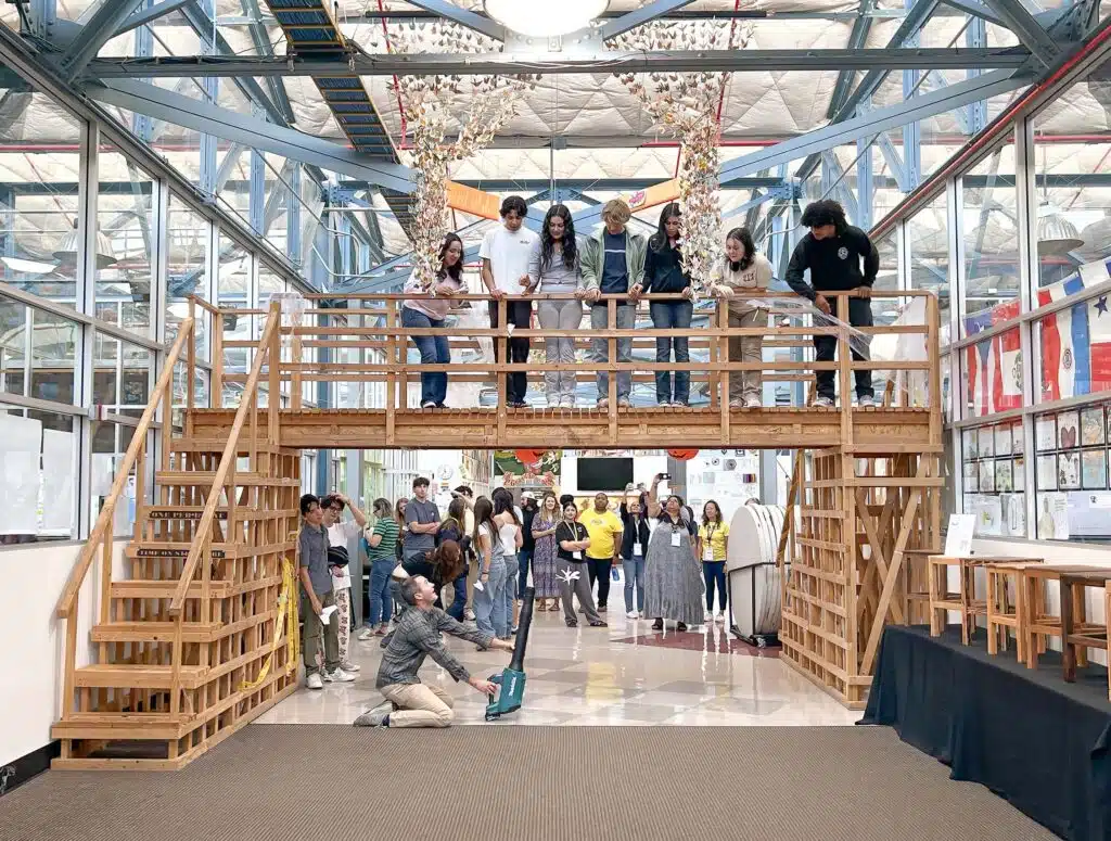 At a leading learning institute, a group tests a wooden bridge indoors. Several individuals stand atop the bridge while others observe from below. A person uses a tool to apply pressure, fostering deeper learning about structural integrity in the bright setting surrounded by various displays.