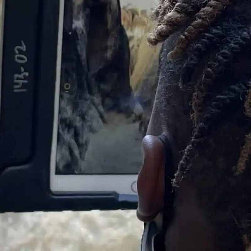 A person with short, twisted hair looks at a tablet showing an image of a rocky canyon or narrow path—perhaps part of a geology project or even a virtual reality geology experience. The side and back of the person’s head are visible in the foreground.