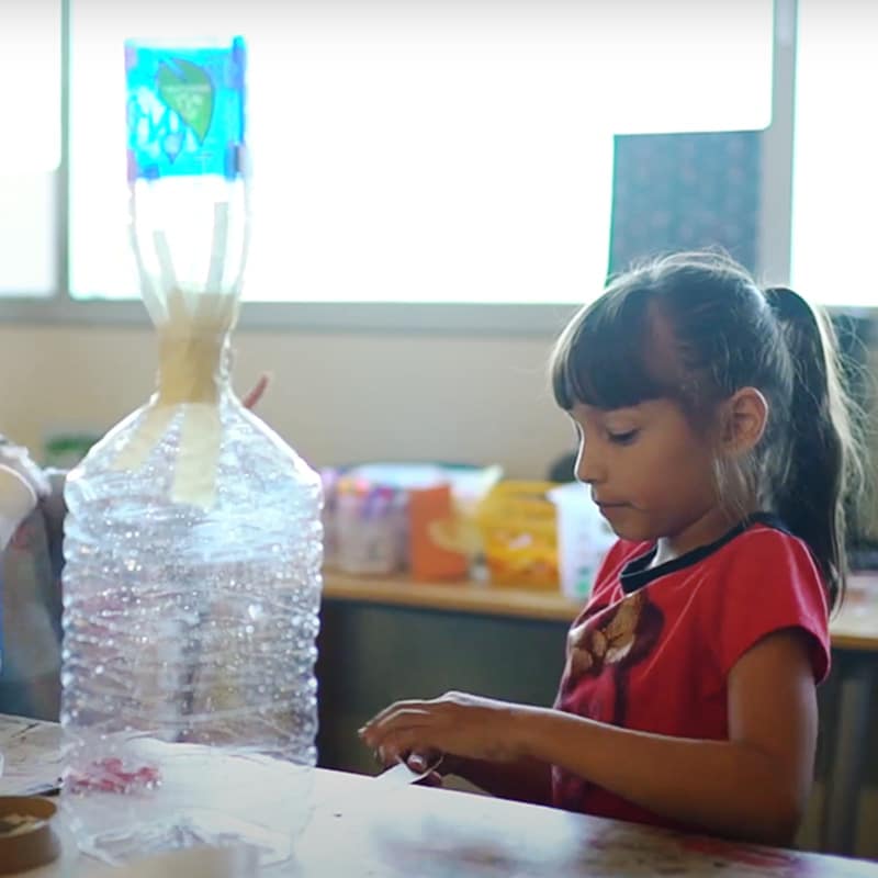 A young girl in a red shirt works on a craft project at a table, with a large plastic bottle structure and art supplies in front of her, showcasing creative student work in a bright, indoor classroom setting.