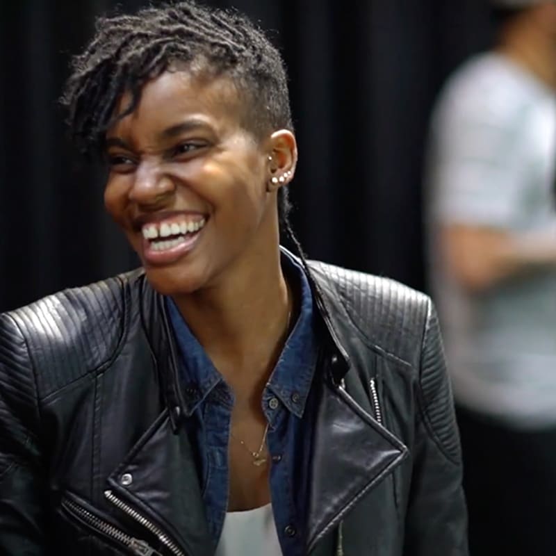 A person with short, styled hair smiles broadly while wearing a black leather jacket over a denim shirt, radiating confidence in knowing your values. The background is blurred with another figure visible.