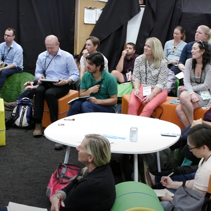 A group of adults sits on colorful furniture around a white table, attentively listening during an indoor meeting or workshop focused on White Fragility Awareness. Some are taking notes, and a can of soda is on the table. The background has black curtains.