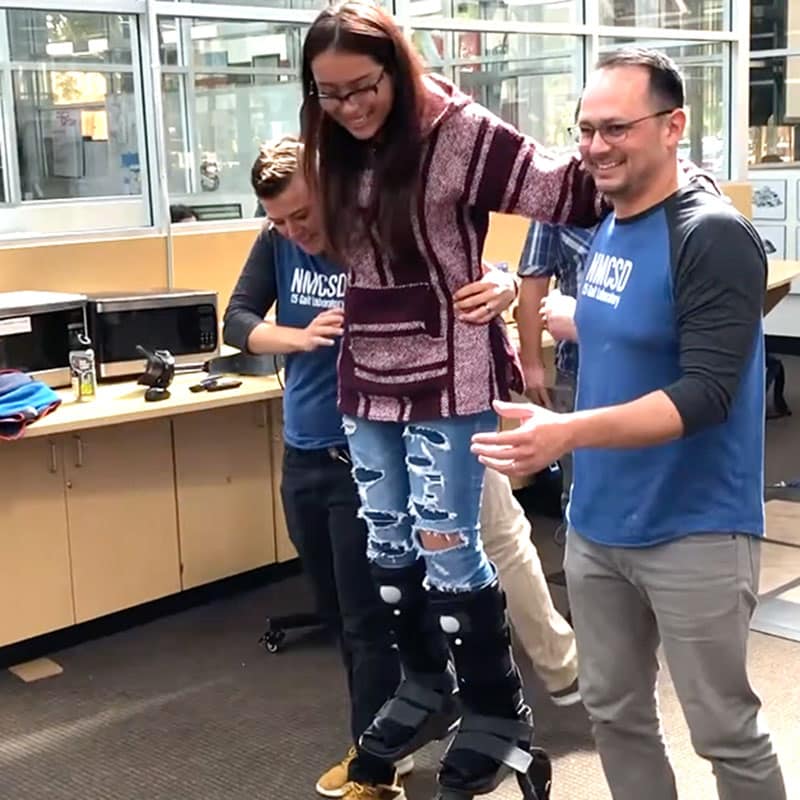 A smiling woman wearing an exoskeleton is supported by two people in a bright indoor space, exemplifying the power of building student strengths, with others watching in the background.