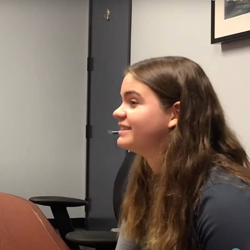 Young woman with long brown hair sits at a table in an office, smiling and looking to the left, perhaps reflecting on her recent internship and valuable work experience. An office chair and closed door are visible in the background, creating a professional setting ideal for career development.
