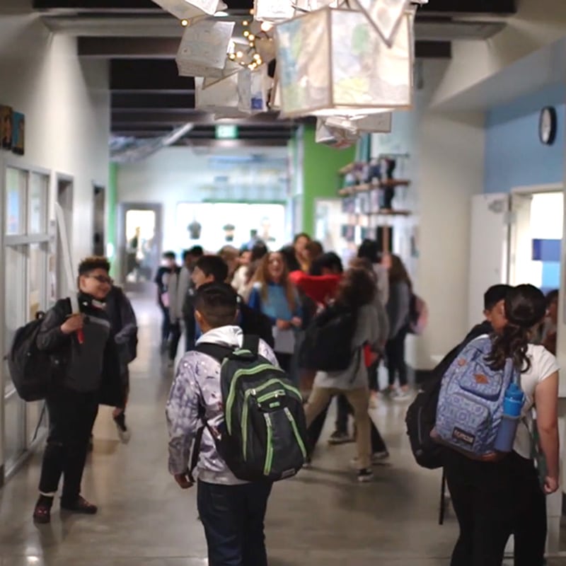 Students with backpacks walk and talk in a brightly lit school hallway, decorated with hanging artwork and colorful walls that reflect the cultural relevance of the diverse student body.