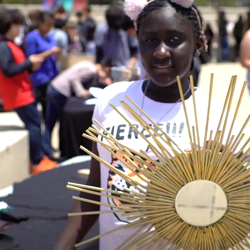 A girl wearing a white shirt holds a round sun-shaped craft with gold rays, embodying high expectations for creativity and achievement. People are gathered at tables outdoors in the background.