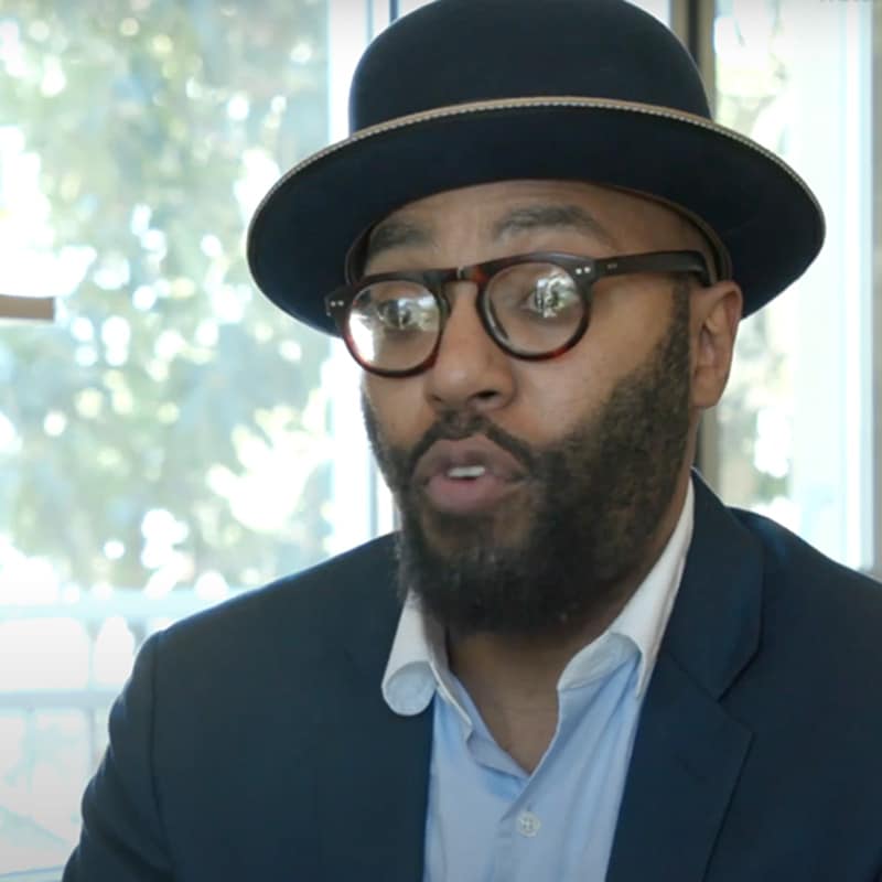 A man with a beard, wearing glasses, a blue suit, and a dark hat with a light band, speaks about online learning in front of a window with blurred greenery outside.