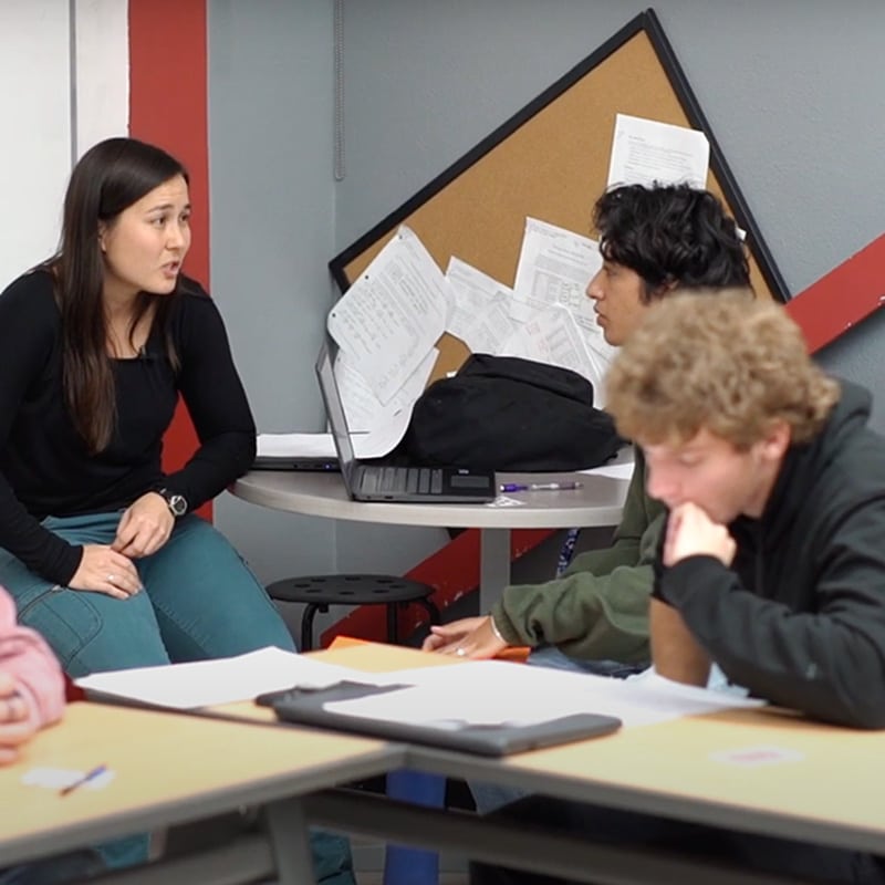 A woman sits at a round table talking to two students, one is listening while another is thinking. Papers and a bulletin board are in the background, suggesting they might be observing schools or discussing their experiences in the classroom environment.
