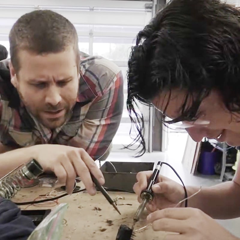 Two people work together at a cluttered table, soldering electronic components. One, embodying teacher mindsets, leans over to offer guidance and support while the other focuses intently on the task. Both are in a workshop setting, collaborating to problem-solve and learn together.