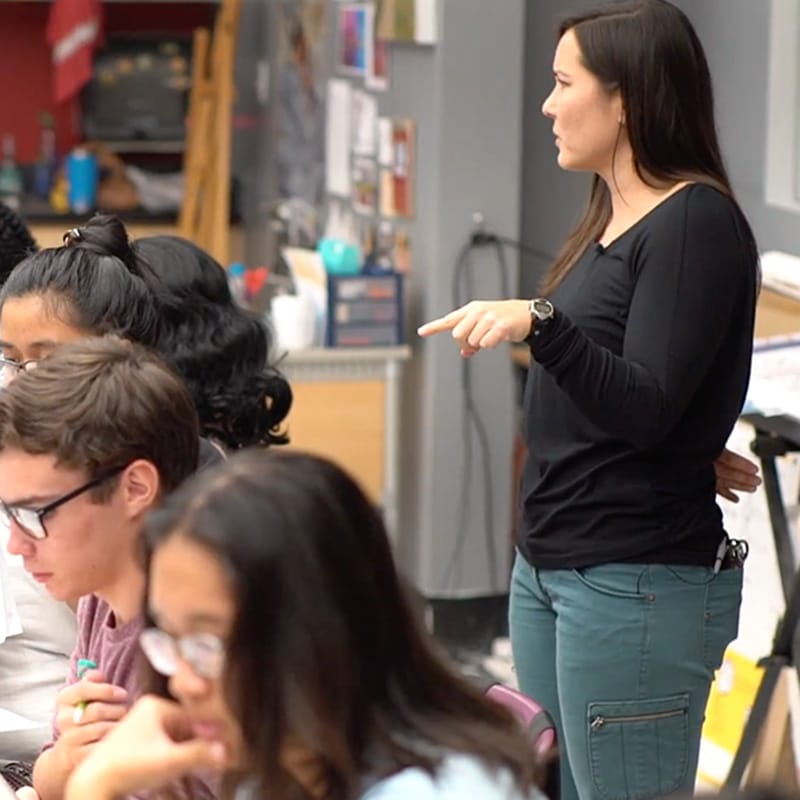 A woman stands and gestures while talking to students seated at a table in a classroom, exemplifying the impact of Teacher Professional Development. The students focus on their work, and classroom materials are visible in the background.