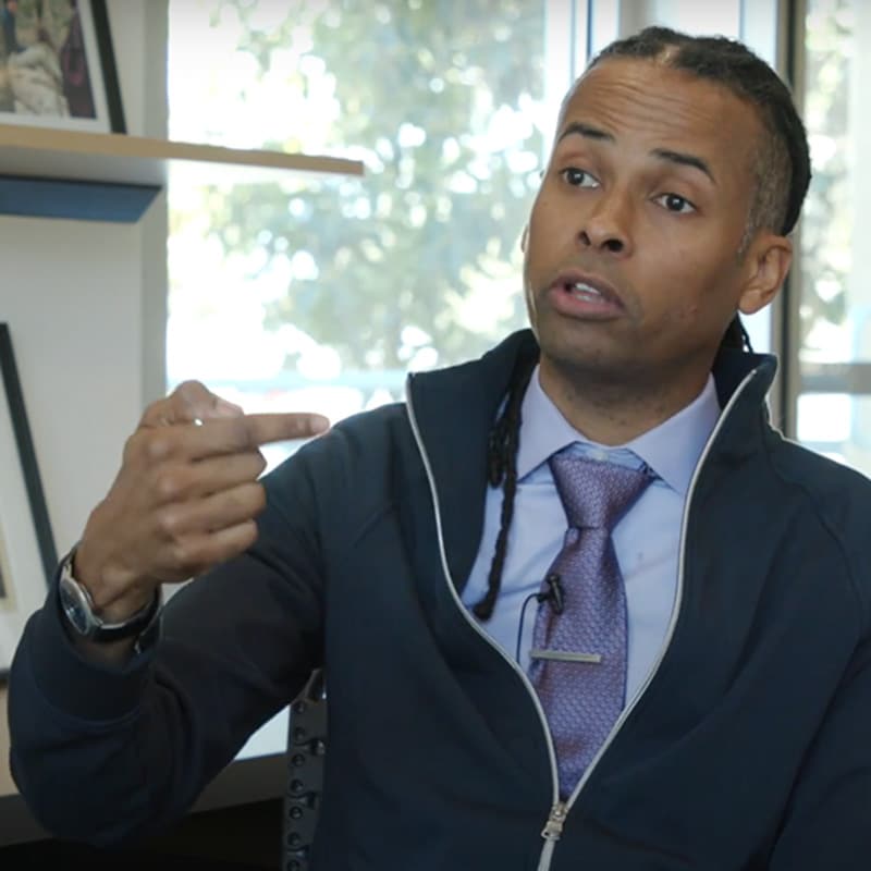 A man in a suit and tie sits indoors, gesturing with his left hand while speaking. As a dedicated learner, he appears to be sharing insights, with shelves of framed photos and a window overlooking trees visible in the background.
