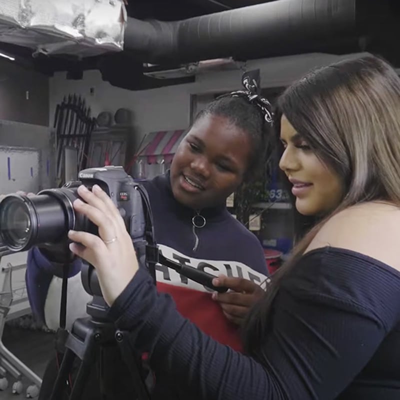 Two women stand together indoors, looking at the screen of a DSLR camera on a tripod, appearing engaged and focused as they review a photo or video in a creative studio setting—a true moment of collaboration that reflects the principles of project-based learning.